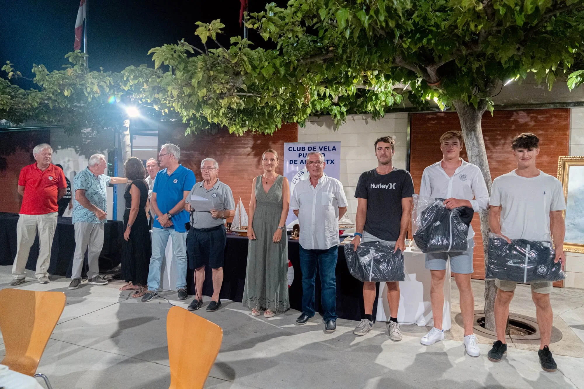 Grupo de personas en una ceremonia al aire libre, algunas sosteniendo premios o bolsas, bajo un árbol en la noche. Hay un cartel que dice 'Club de Vela Puerto de Alicante'.