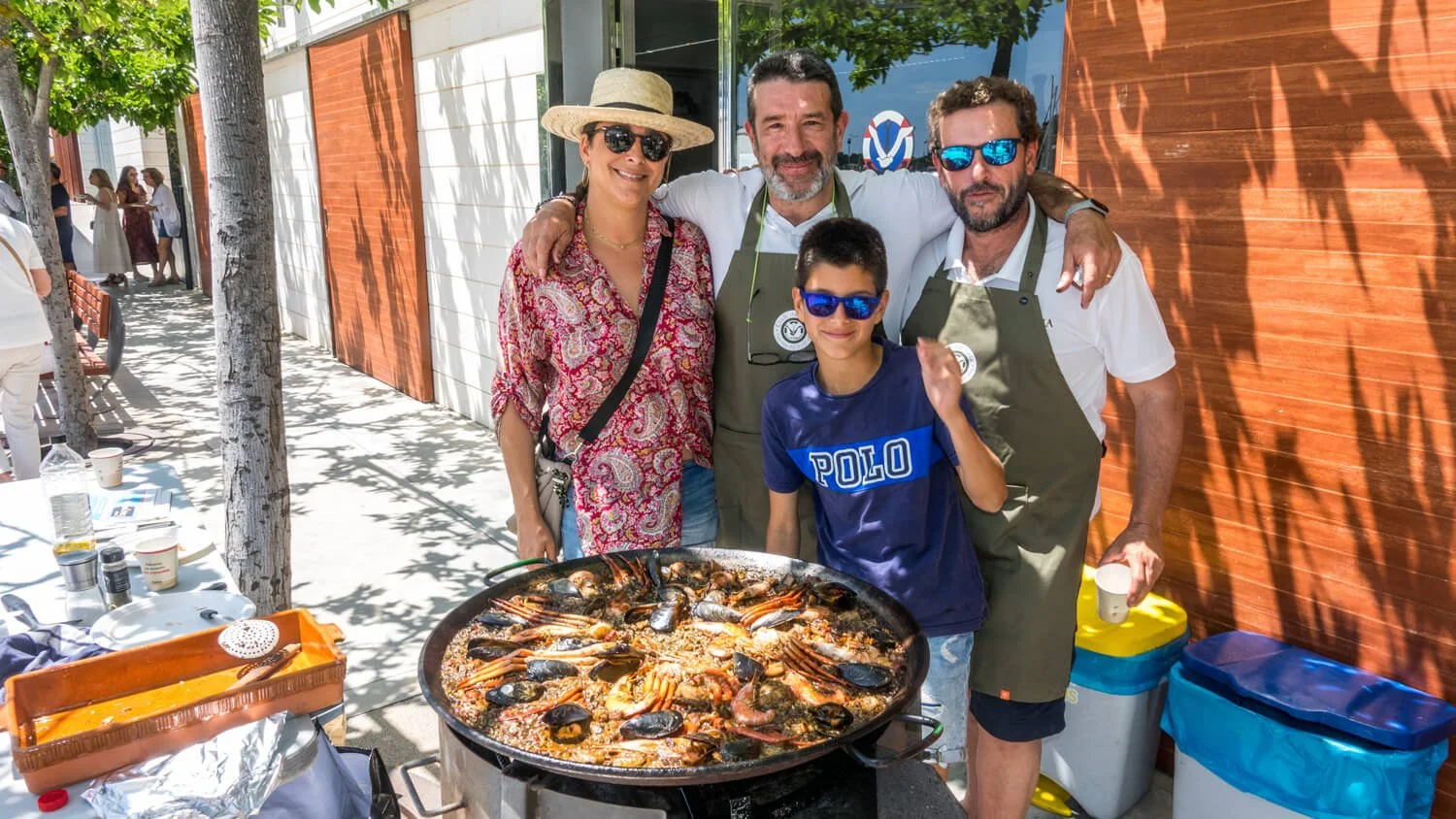 Grupo de cuatro personas sonriendo, rodeadas de sombra de árboles, en una comida exterior con un plato grande de mariscos y arroz en primer plano.