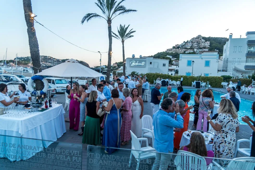 Gente en una fiesta al aire libre cerca de una piscina, con mesas, comidas, bebidas y palmeras, en un ambiente ameno en un entorno costero con colinas y edificaciones blancas en el fondo.