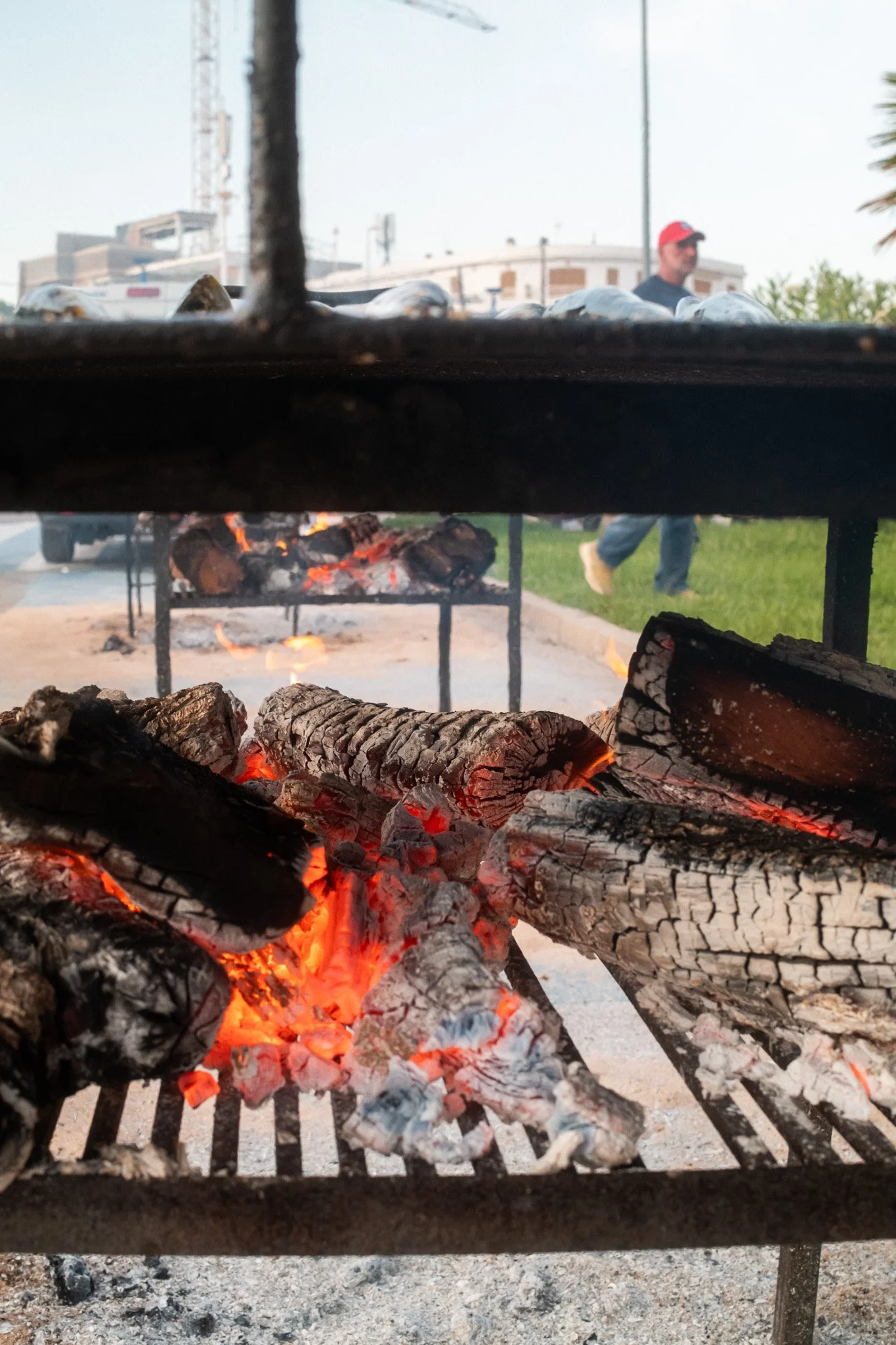 Carbón encendido sobre una parrilla de barbacoa vista desde abajo, con una persona caminando en el fondo, en un espacio exterior.