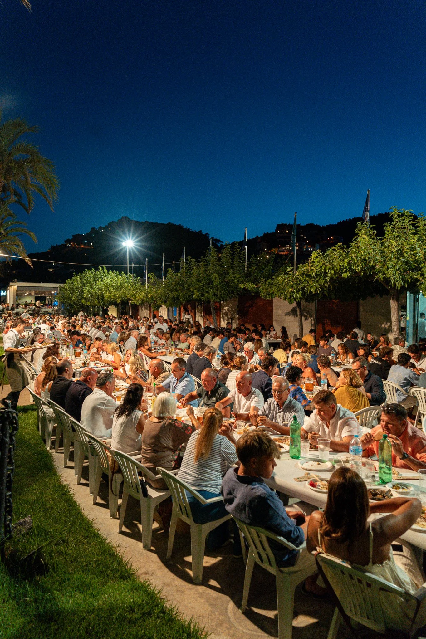 Una gran reunión al aire libre con muchas personas sentadas en mesas, cenando durante la noche, con árboles y montañas al fondo y un cielo oscuro con algunas estrellas.