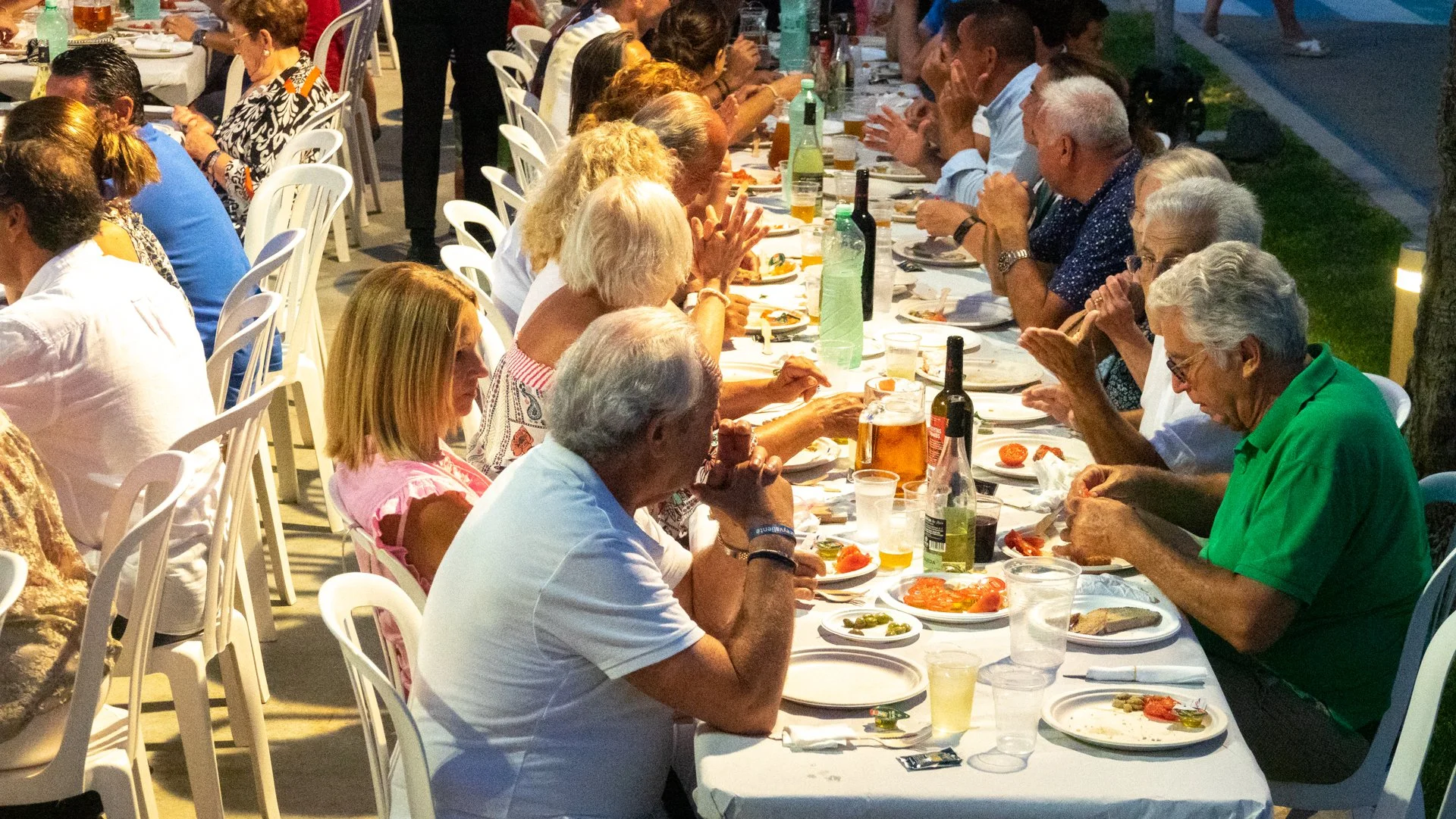 Personas sentadas en una larga mesa al aire libre durante la noche, comiendo y bebiendo juntos en una reunión social.