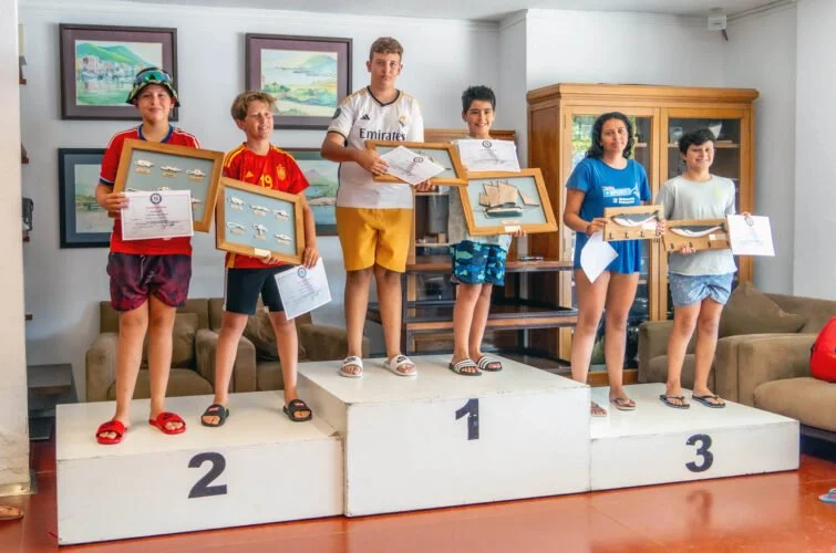 Niños en un podio de premiación, sosteniendo premios y certificados en un ambiente interior con decoraciones de cuadros y muebles.