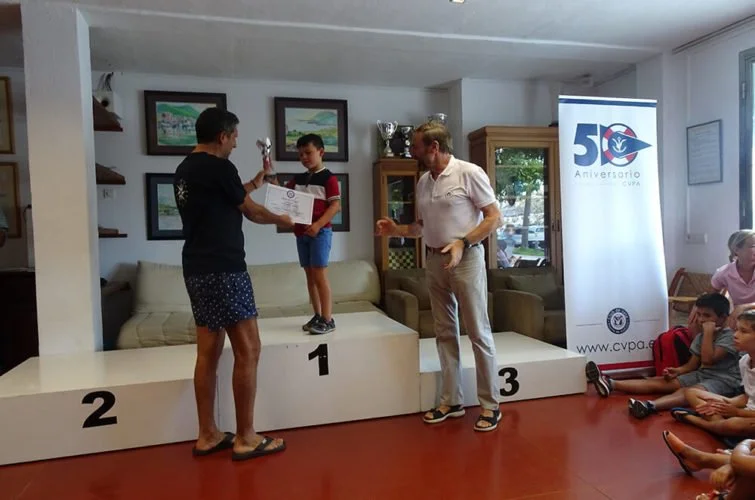 Un niño en el primer lugar de una ceremonia de premiación recibe un trofeo de un hombre mientras un adulto se acerca y otros niños observan sentados en el suelo, en un ambiente interior con decoraciones y un banner de celebración.