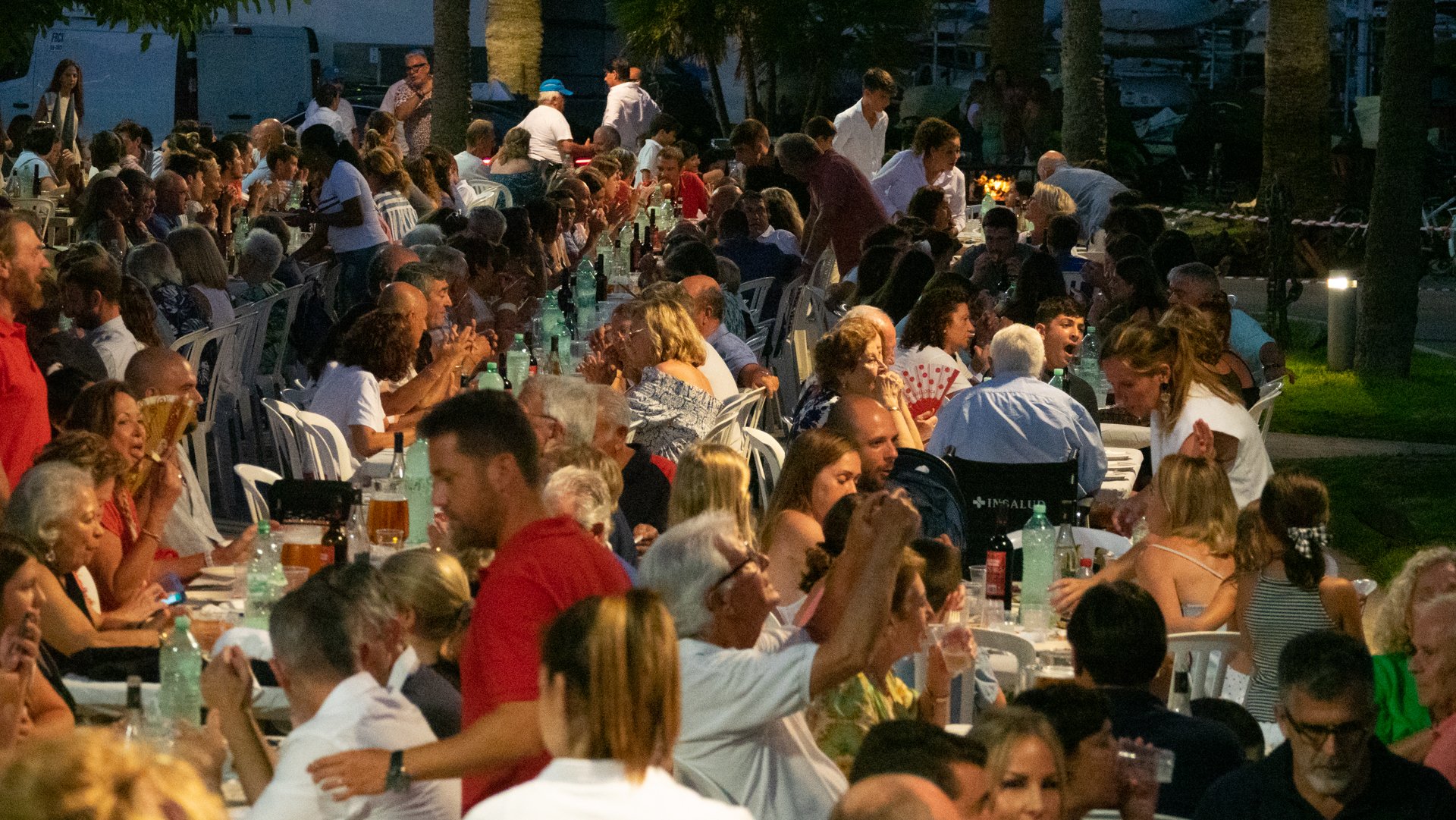 Cena al aire libre con muchas personas sentadas en mesas largas, cenando y conversando, en un ambiente nocturno con árboles y luces suaves.