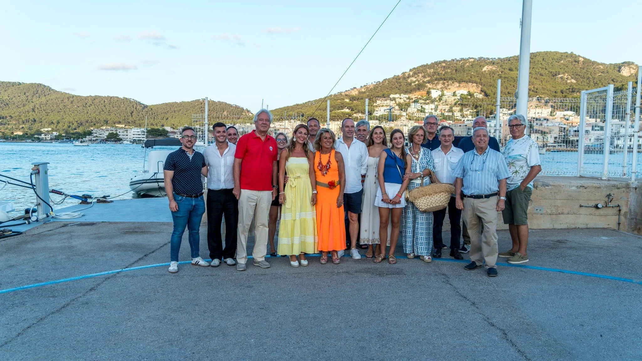 Grupo de personas posando frente a un muelle con montañas y casas en el fondo, en un día soleado.