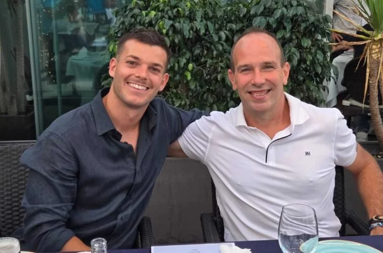 Dos hombres sonriendo y sentados juntos en una mesa al aire libre en un restaurante o cafetería.