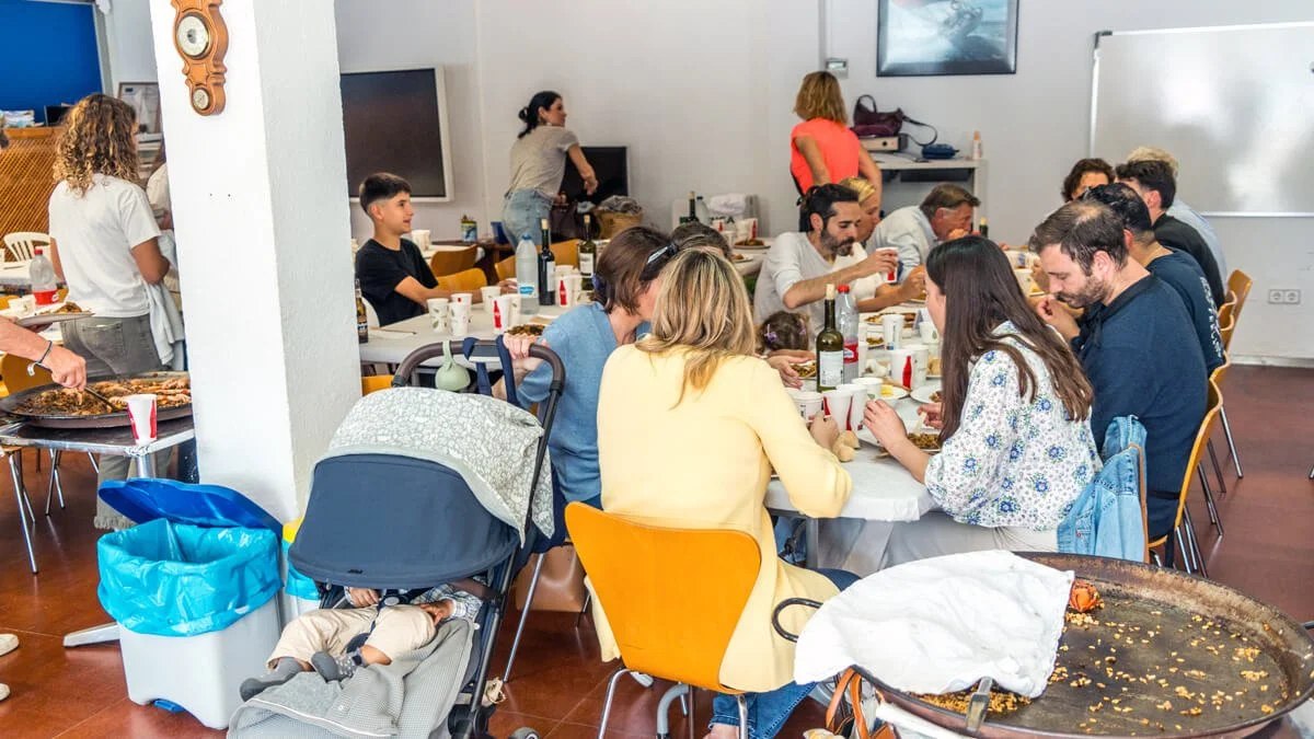 Personas comiendo en una comida comunitaria en un salón, con varias mesas y comida servida; en primer plano, una carreola con un bebé durmiendo