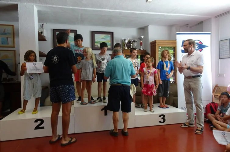 Niños en podio en un evento de premiación en una sala con trofeos y cuadros en las paredes, adultos entregando premios y tomando fotos.