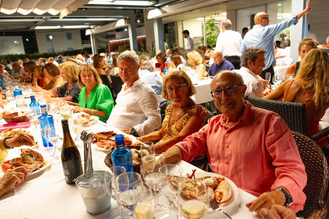 Grupo de personas celebrando en una fiesta o reunión al aire libre, algunas con comida y bebida en una mesa larga