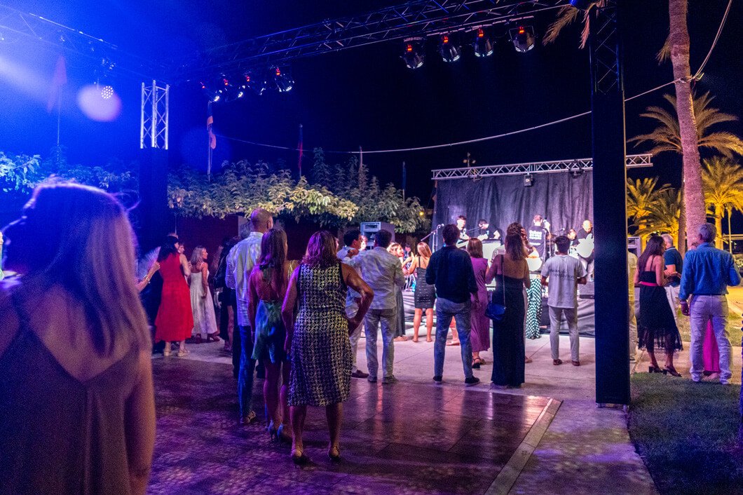 Gente bailando y disfrutando de un concierto al aire libre por la noche, con escenario y músicos en el fondo, rodeados de palmeras y vegetación.