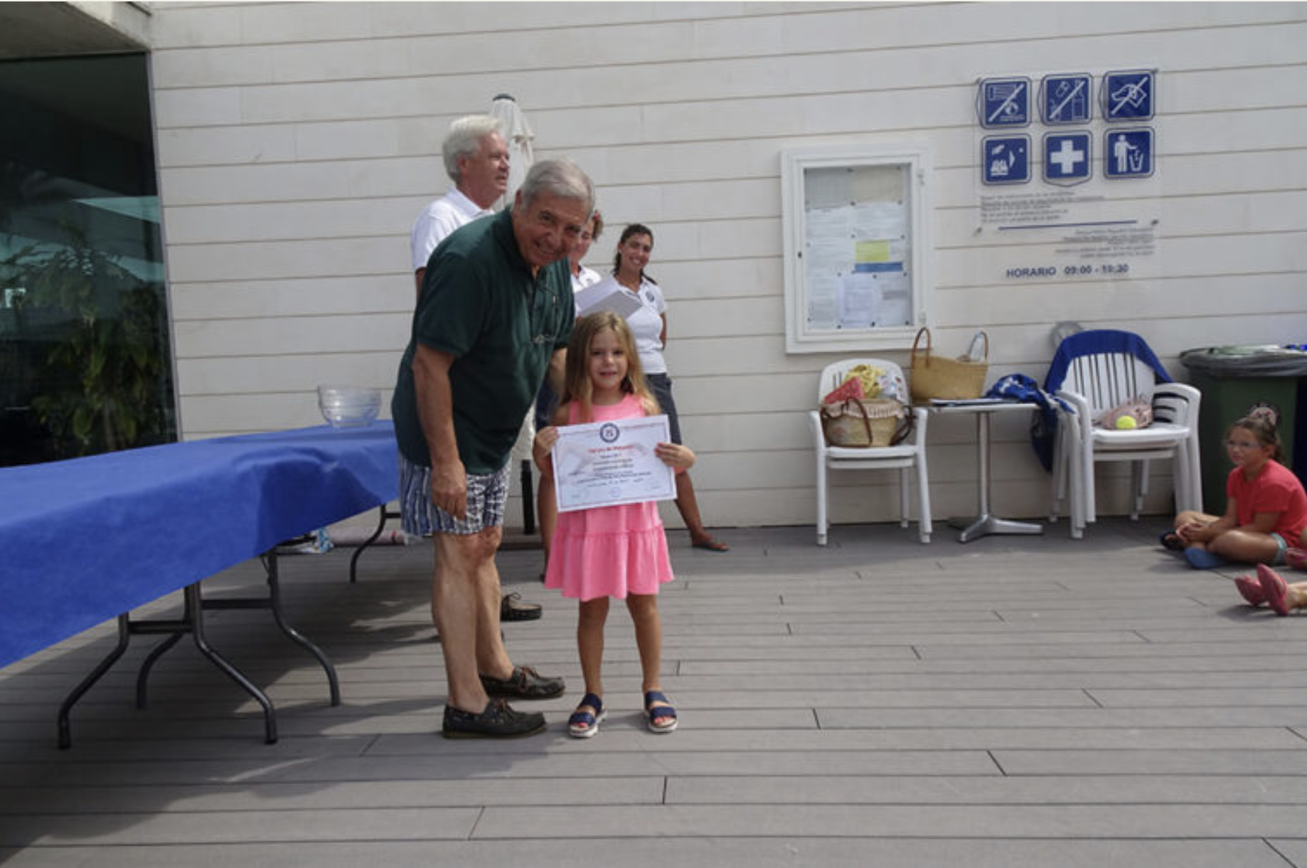 Niña recibe un certificado de reconocimiento durante ceremonia en un espacio abierto con compañeros y adultos observando, mesas y sillas al fondo.