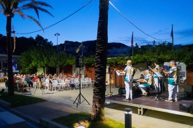 Conjunto musical tocando en un escenario al aire libre en la noche, mientras una audiencia observa sentada en sillas, con árboles y edificios en el fondo.