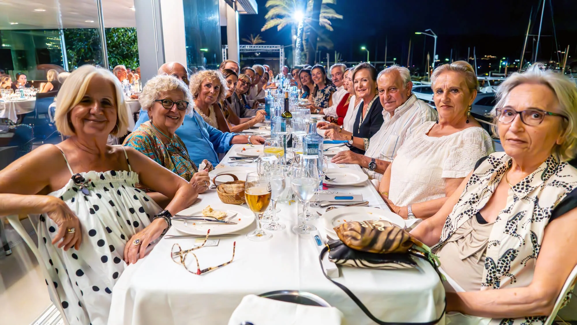 Grupo de personas mayores sentadas en una mesa en un restaurante al aire libre por la noche, celebrando una reunión social con comida y bebidas, en un ambiente animado y alegre.