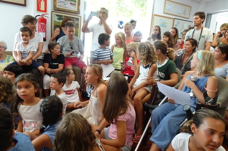 Grupo de niños y adultos en una sala, algunos sentados y otros de pie, participando en una actividad o evento