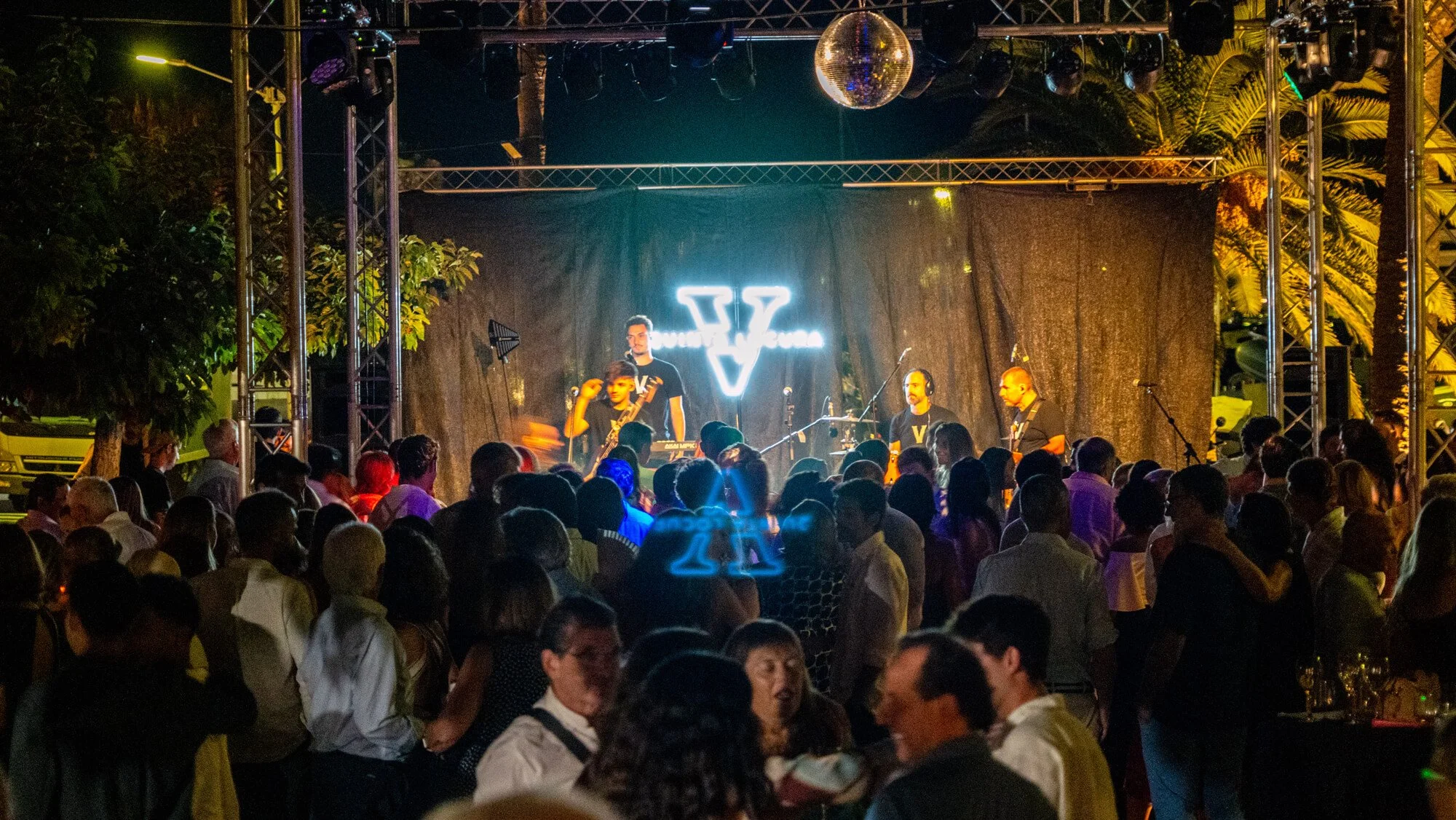 Concierto en vivo al aire libre con músicos en un escenario decorado con luces y una bola disco, rodeado por una multitud de espectadores en la noche.