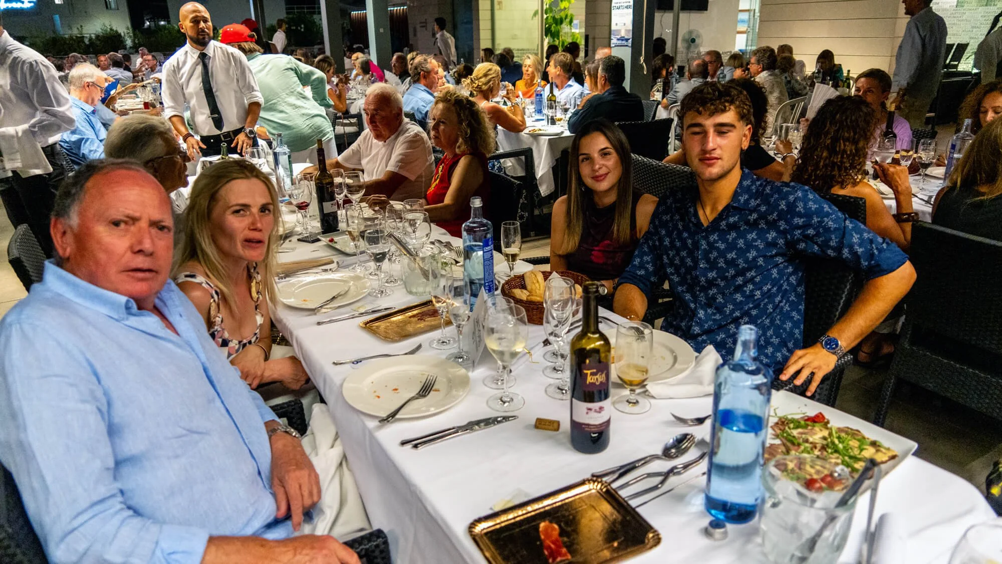 Personas sentadas en una mesa de restaurante durante una celebración, con comida, vino y decoraciones en un ambiente luminoso y concurrido.