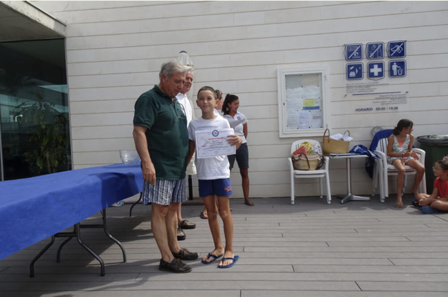 Un grupo de niños y adultos en una ceremonia de premiación al aire libre, en un lugar con paredes de madera blanca y un cartel con símbolos de servicios de salud y horario.