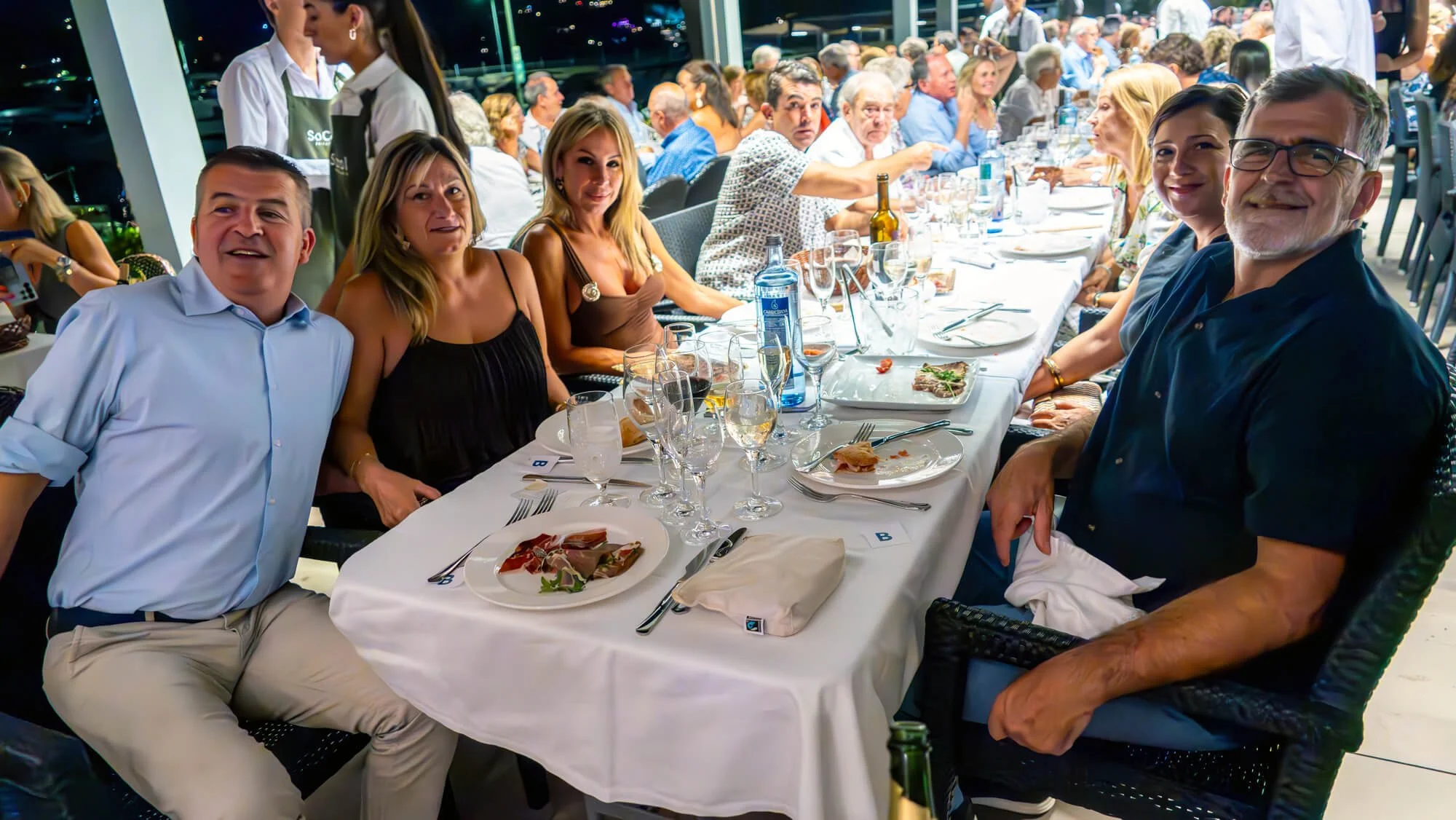 Grupo de personas sentadas en una mesa durante una cena en un ambiente sofisticado, algunos sonriendo y otros conversando, con platos de comida y vasos con bebida en la mesa.