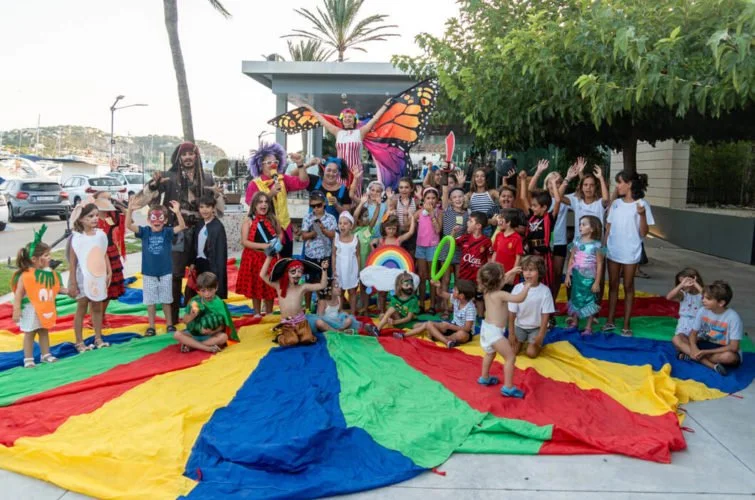 Grupo de niños y adultos en un evento al aire libre con un payaso y una mariposa gigante, jugando sobre un paracaídas colorido.