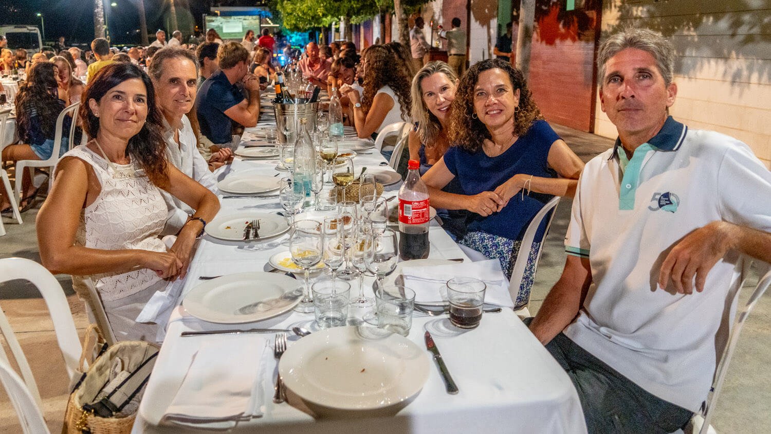 Grupo de personas en una cena al aire libre en una reunión o celebración, con mesas decoradas con utensilios y bebidas, en un ambiente nocturno y festivo.
