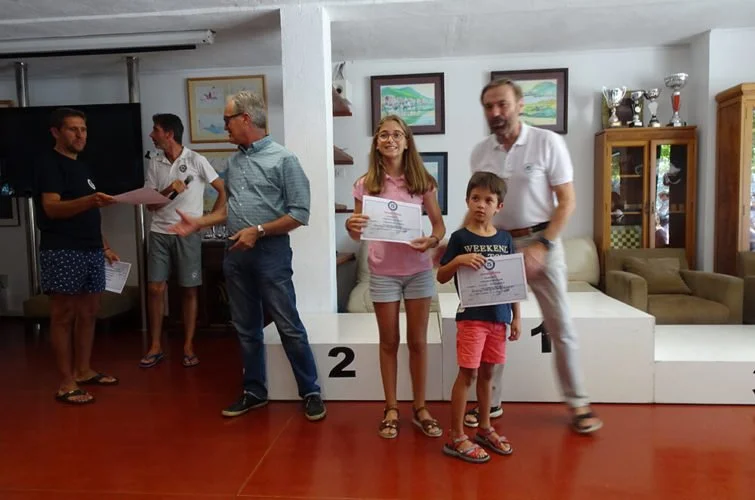 Niña y niño sosteniendo certificados en un escenario de premiación, con otros adultos en el fondo en una sala decorada con cuadros y trofeos.