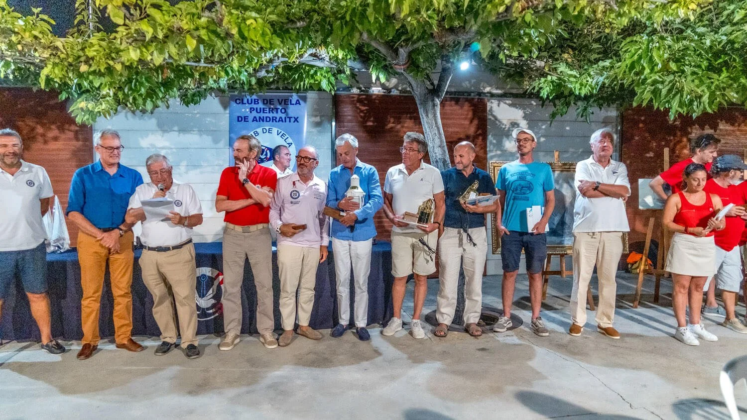 Grupo de personas en una ceremonia de premiación, con algunos sosteniendo trofeos, en un entorno al aire libre con árboles y una pared de fondo. 