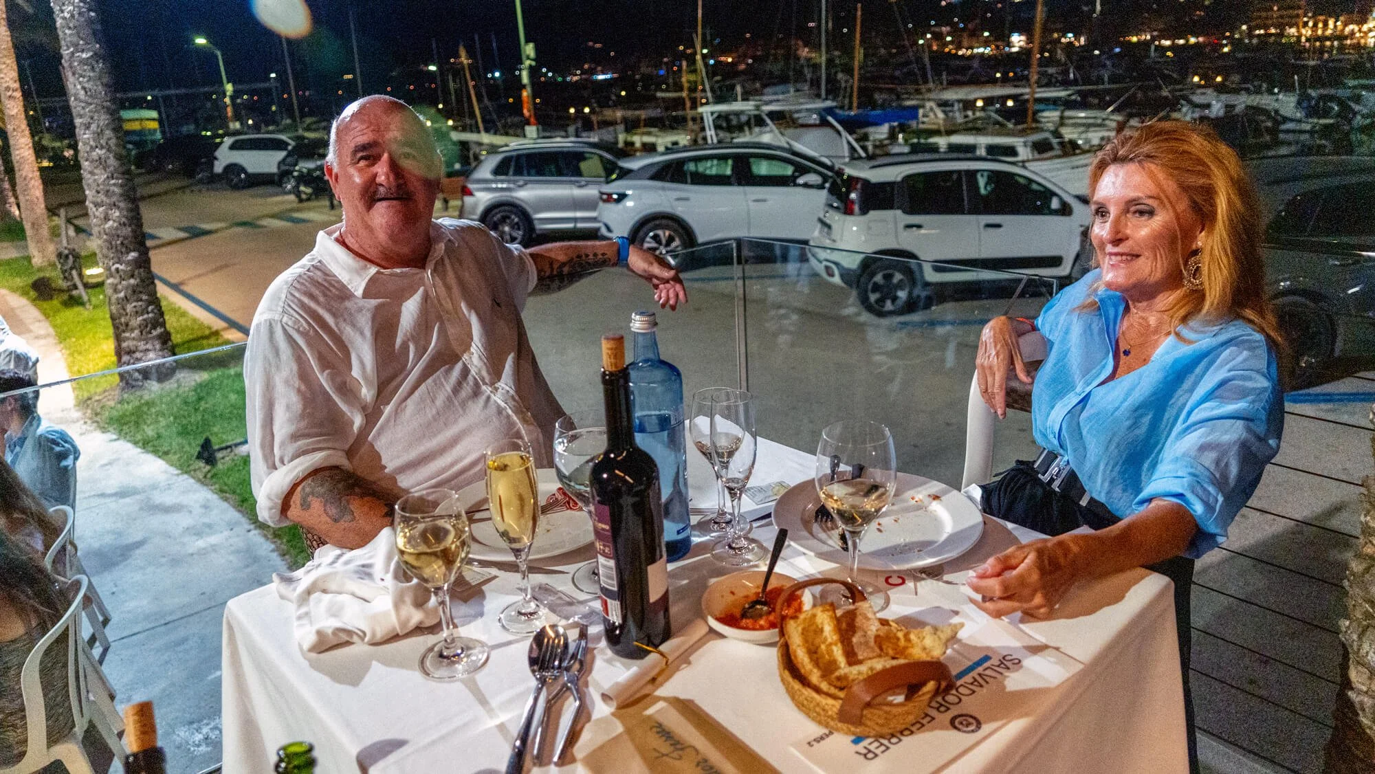 Pareja sentada en una mesa de restaurante al aire libre con platos de comida, vino y bebidas, en la noche, con un fondo de marina con barcos y automóviles estacionados.