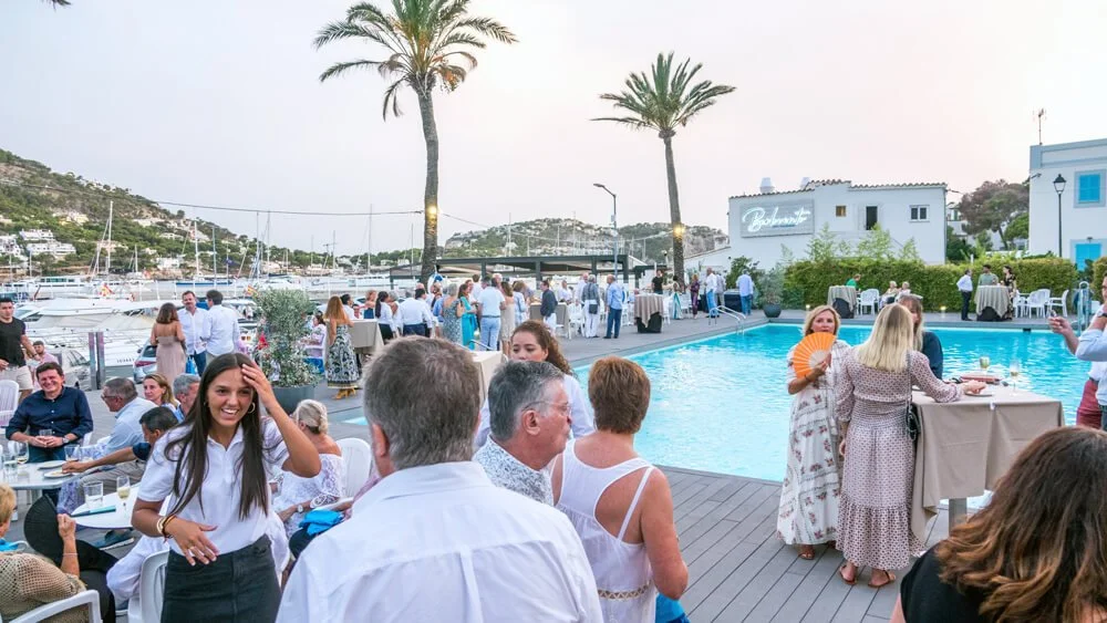 Celebración al aire libre junto a una piscina, con muchas personas socializando y disfrutando, en un lugar con vista al mar y palmeras, durante el atardecer.