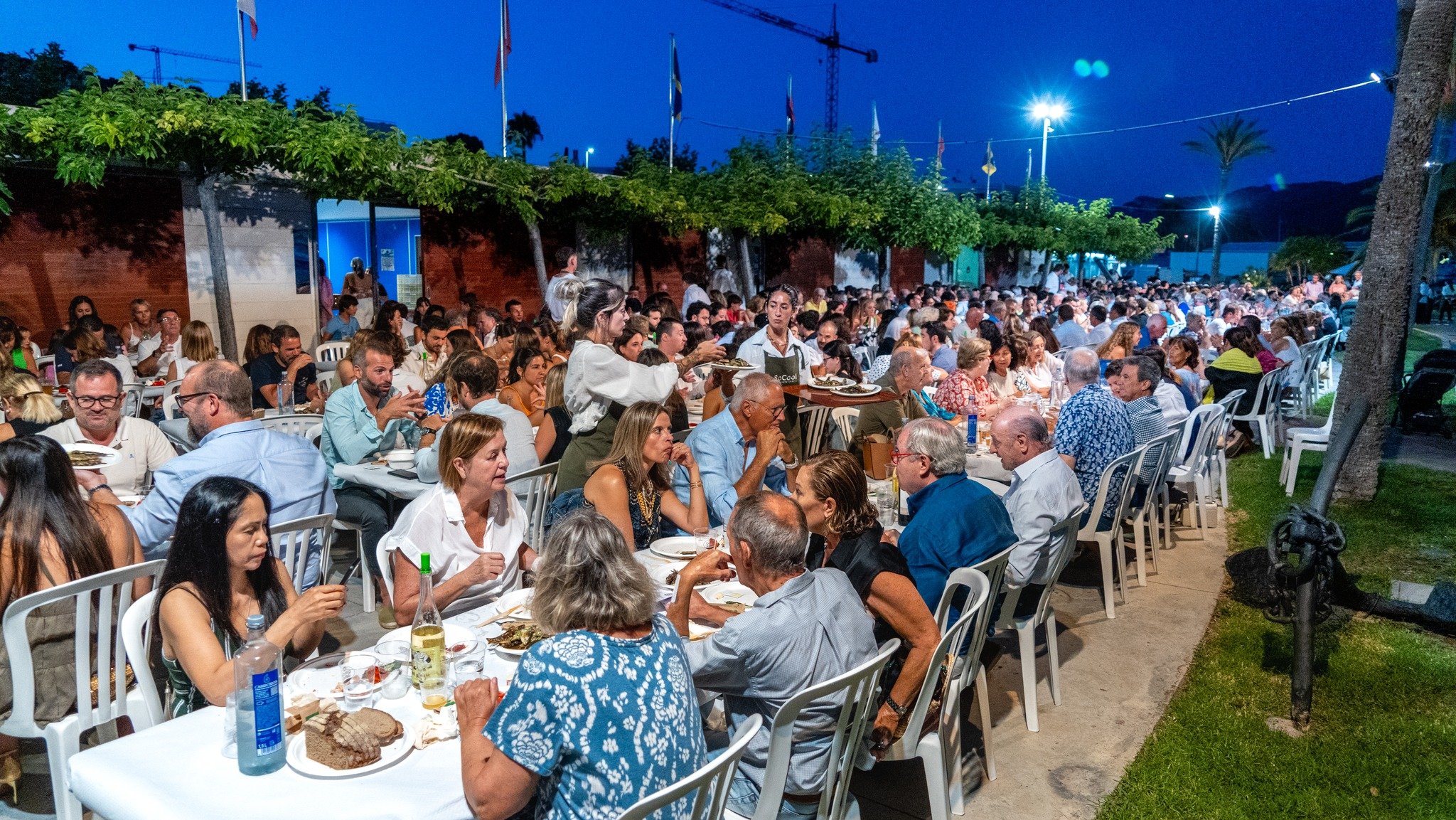 Una gran reunión al aire libre con muchas personas sentadas en mesas, disfrutando de la comida en una fiesta nocturna, con árboles, banderas y estructura de construcción en el fondo.