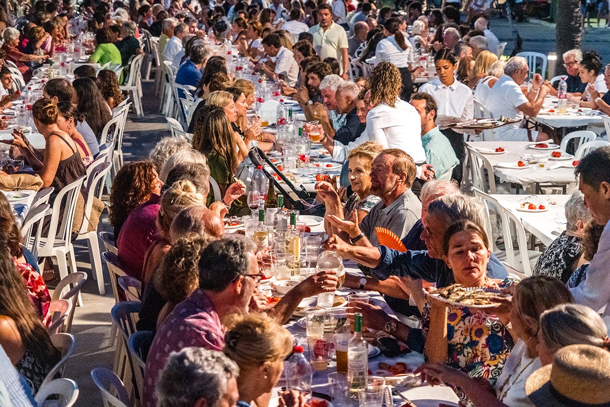 Una gran mesa con muchas personas comiendo y bebiendo en un evento al aire libre durante el día.