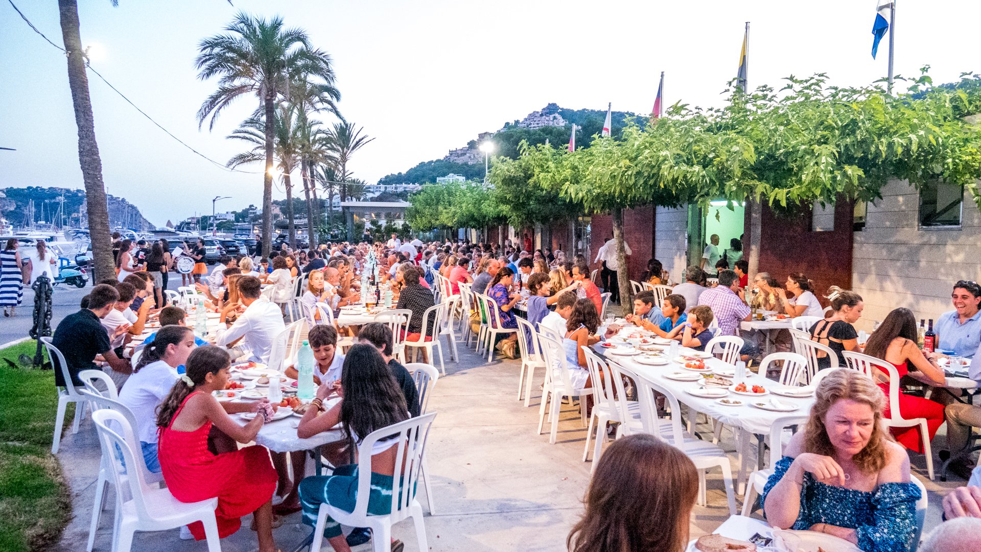 Escena de una cena al aire libre con muchas personas sentadas en mesas largas, sobre un paseo junto a un puerto con yates y montañas de fondo, en una tarde o noche.