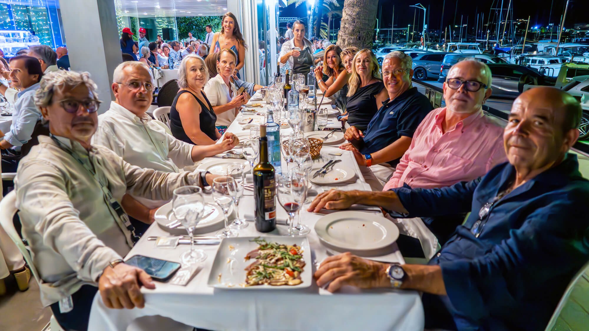 Grupo de personas sentadas en una mesa de restaurante durante la noche, con comida y bebidas, en un ambiente elegante con vista de un puerto con lanchas y una terraza con luces