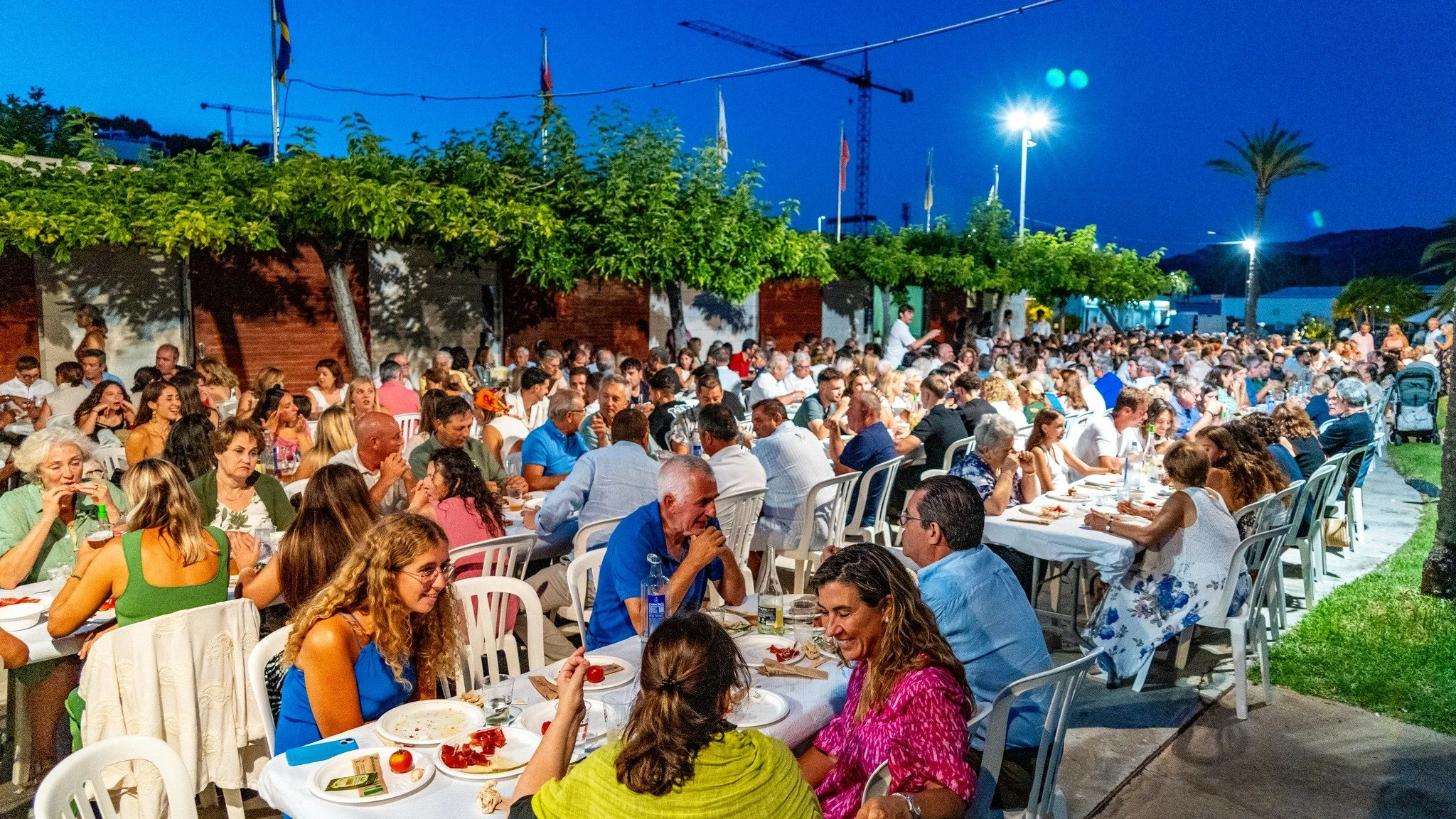 Una gran reunión al aire libre por la noche con muchas personas sentadas en mesas en un lugar decorado con árboles. Hay luces y algunas banderas en el fondo.