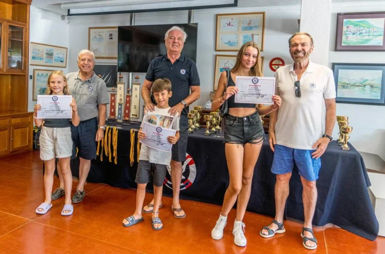 Grupo de seis personas, incluyendo niños y adultos, posando con diplomas y trofeos en una sala con cuadros en las paredes y un televisor grande.
