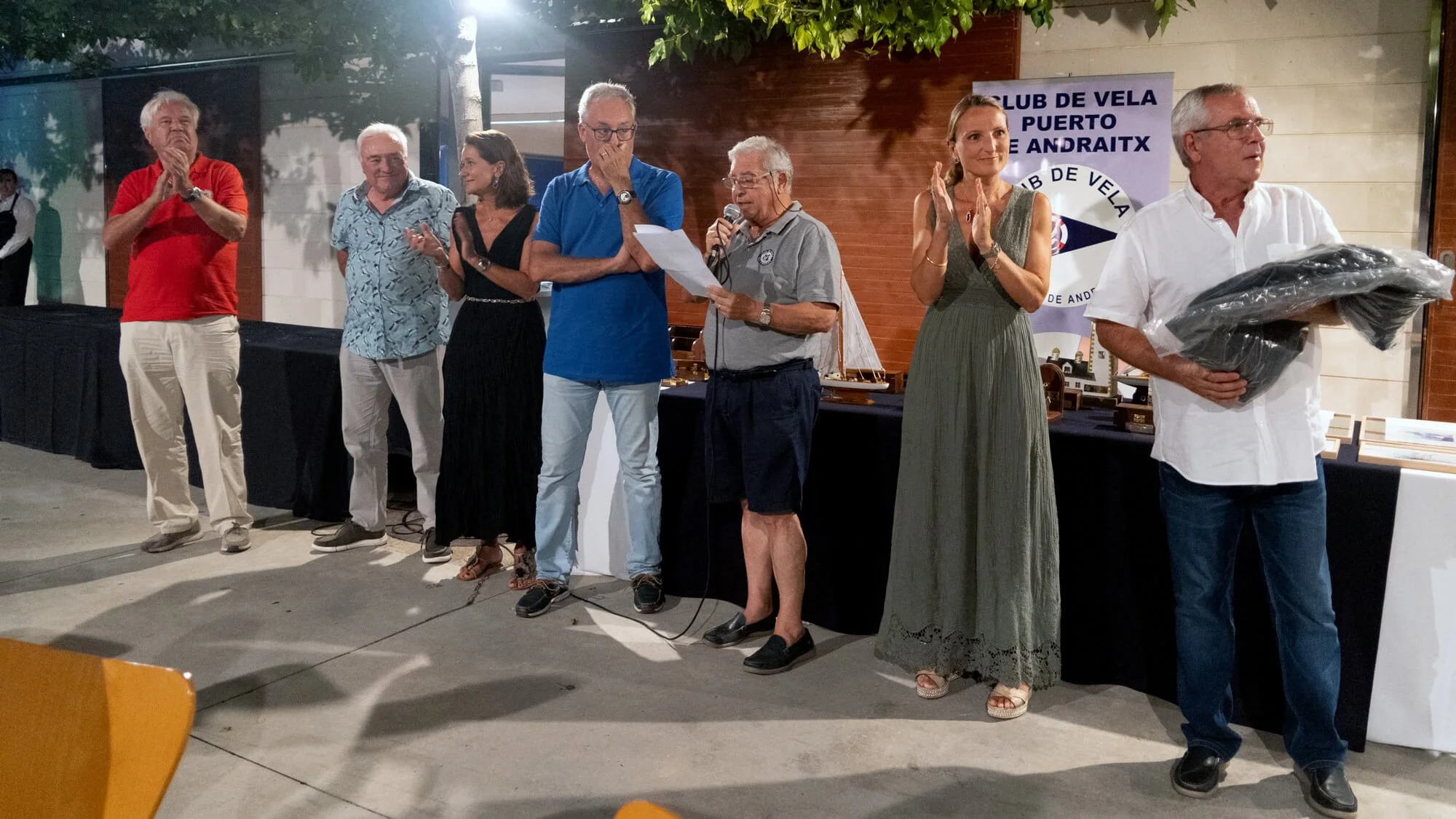 Grupo de personas en una ceremonia o evento, algunas aplaudiendo y una leyendo un discurso, en un espacio cerrado con decoración de madera y un banner del club de vela 'Puerto Andraitx' en el fondo.