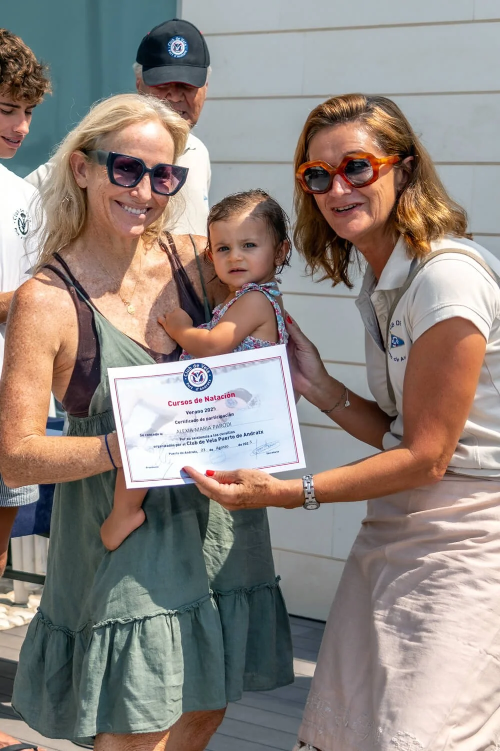 Mujer mayor con gafas de sol recibiendo un certificado de participación en cursos de natación, acompañada de una niña y otra mujer en un acto de entrega de reconocimientos al aire libre.