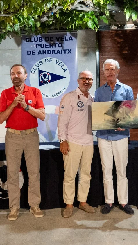 Tres hombres de pie en una ceremonia de reconocimiento, con un banner de un club de vela en el fondo, uno de ellos sosteniendo una pintura o trofeo, bajo un toldo con follaje.