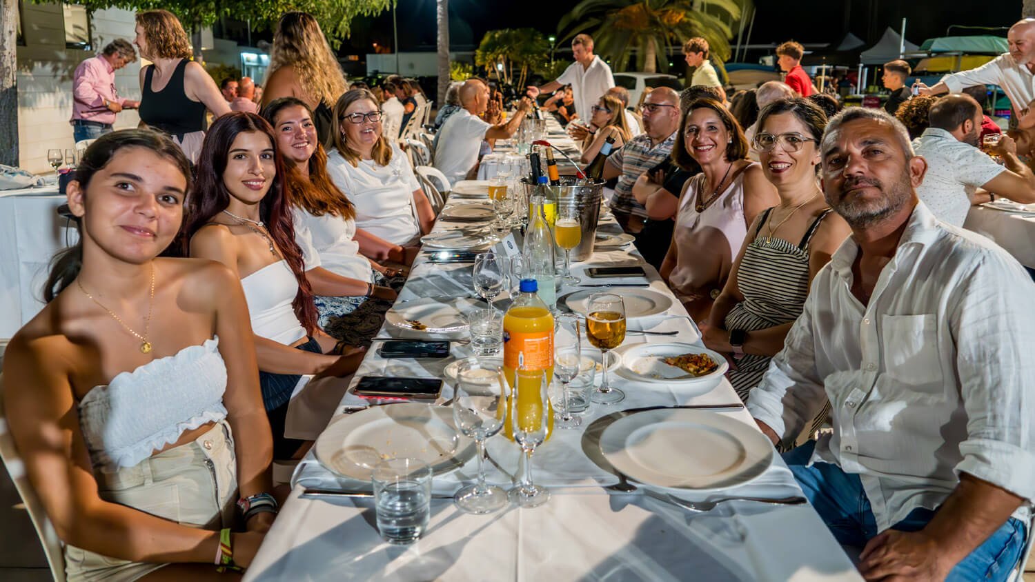 Grupo de personas cenando en una mesa larga en un evento al aire libre durante la noche, con comida y bebidas en la mesa.