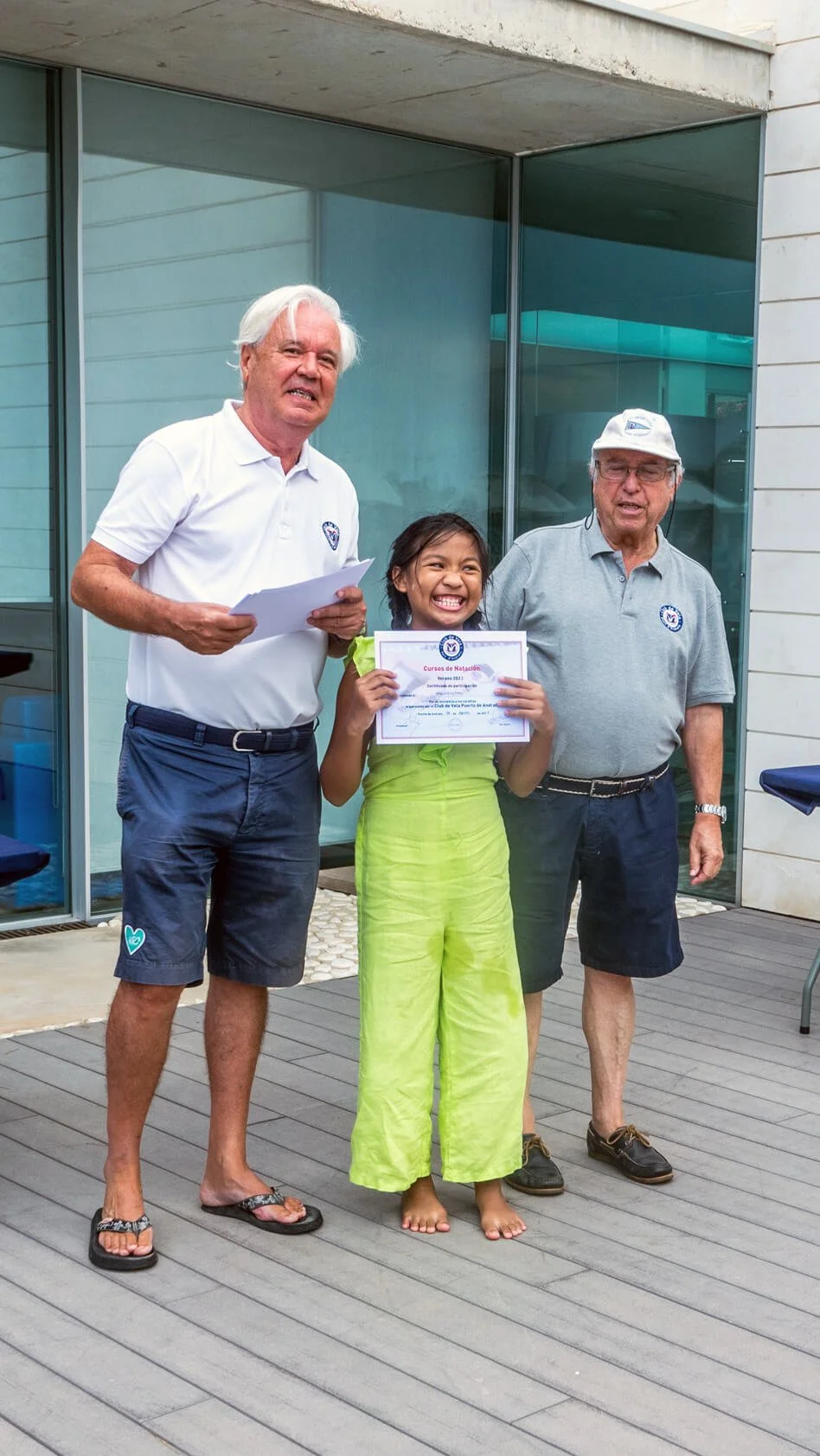 Una niña recibe un diploma de natación en una ceremonia, acompañada por dos hombres mayores, uno de ellos sosteniendo papeles; todos están sonriendo y de pie en un área exterior con suelo de madera y fondo de paredes de vidrio.