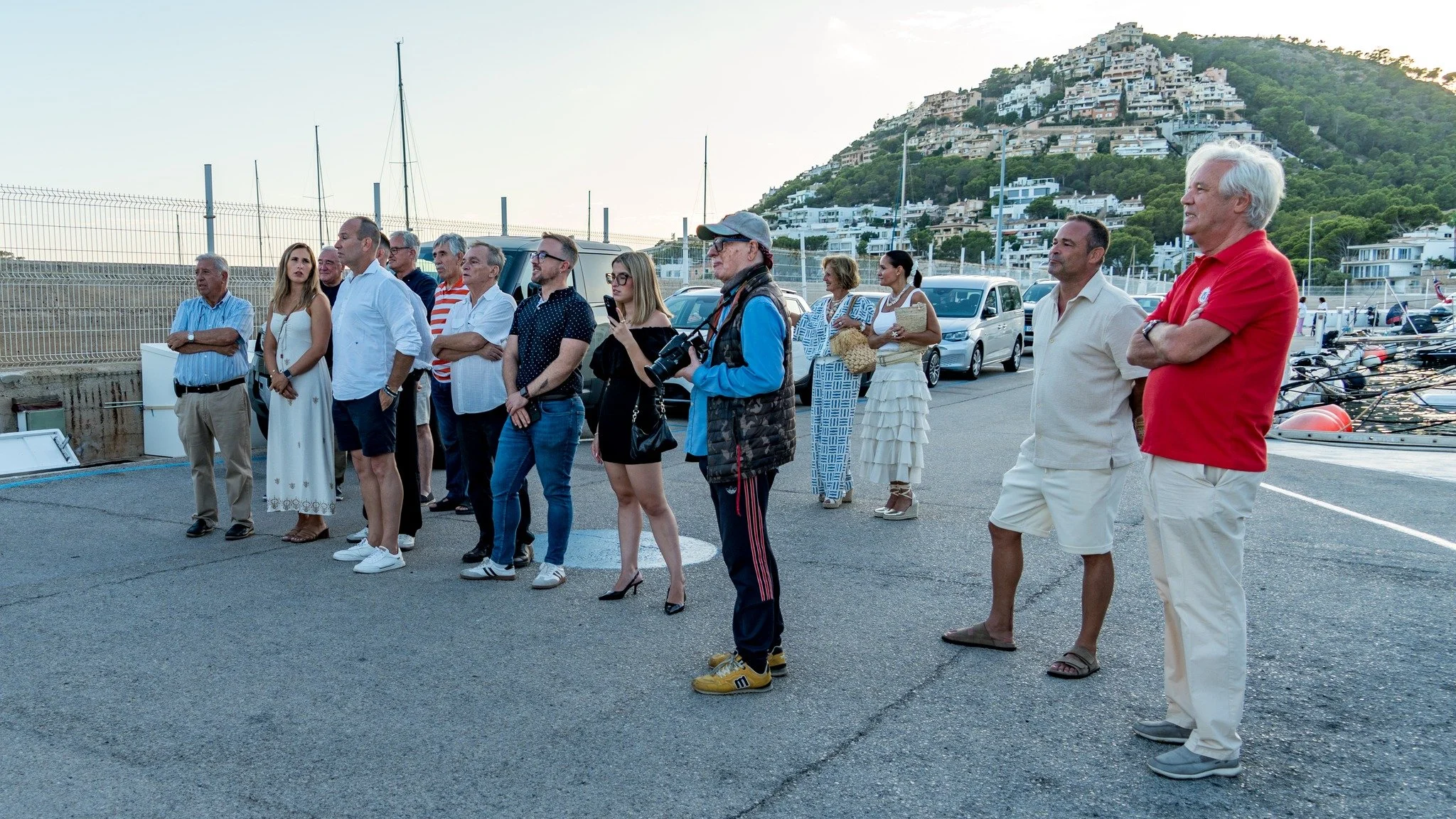 Grupo de personas observando algo en un muelle cerca de la playa con colinas con casas blancas en el fondo.