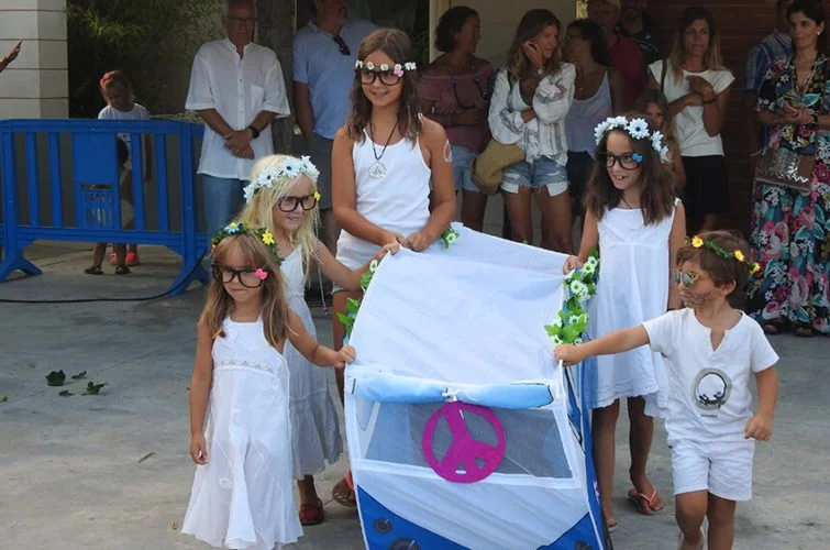 Niñas vestidas con ropa blanca, algunas con coronas de flores, participando en una marcha pacífica con un carrito decorado con flores y símbolos de paz, en un entorno al aire libre con varias personas observando.
