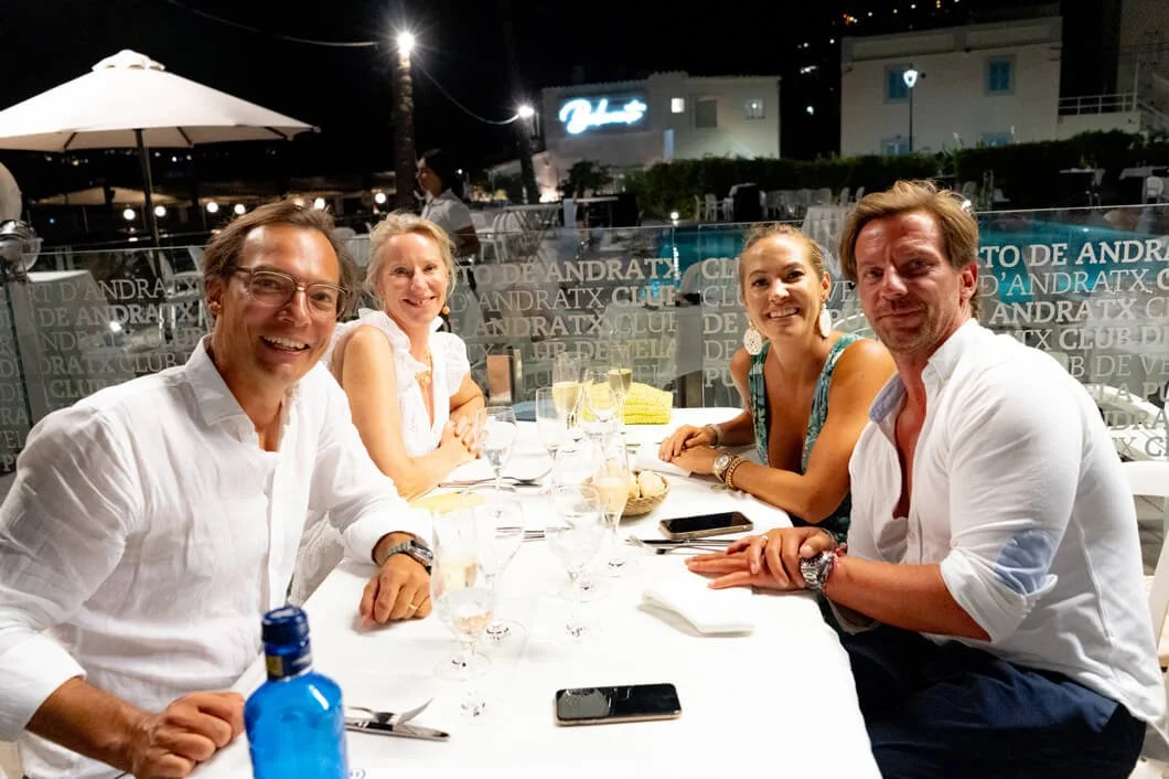 Cinco personas sentadas en una mesa en un restaurante al aire libre en la noche, disfrutando de una comida y bebidas, con fondo de luces y edificios.
