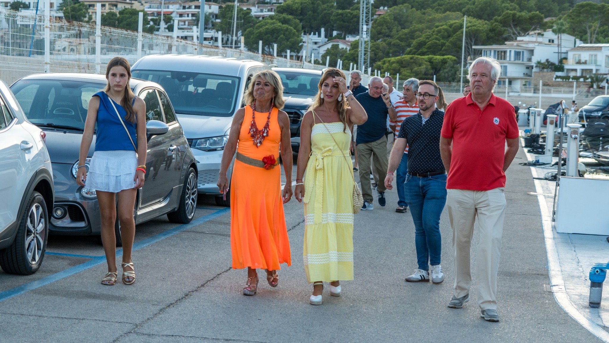 Grupo de personas caminando por una zona de estacionamiento, algunas vestidas con ropa de colores vivos, junto a autos y en un entorno con colinas y casas.
