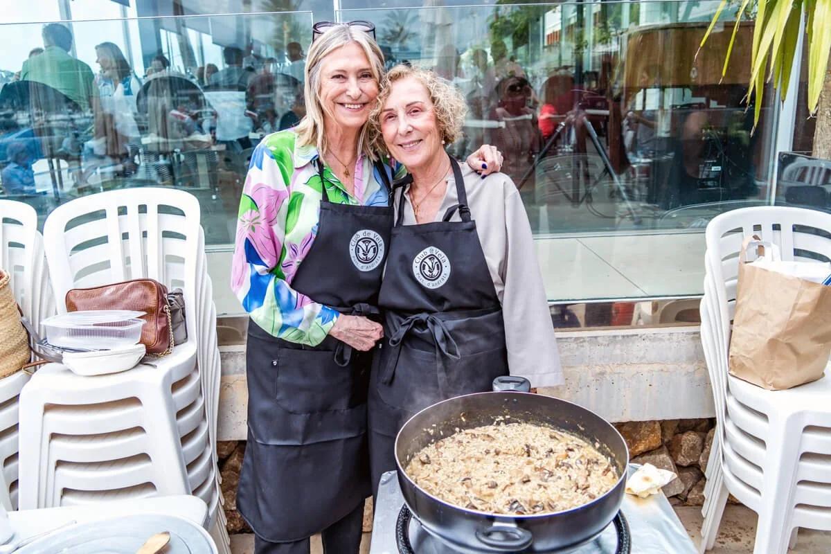 Dos mujeres mayores sonriendo en una cocina, vestidas con delantales negros, una de ellas con una blusa colorida. Están cocinando en una sartén grande con un guiso, cerca de un ventanal con vista a una gente en el interior de un lugar cerrado.