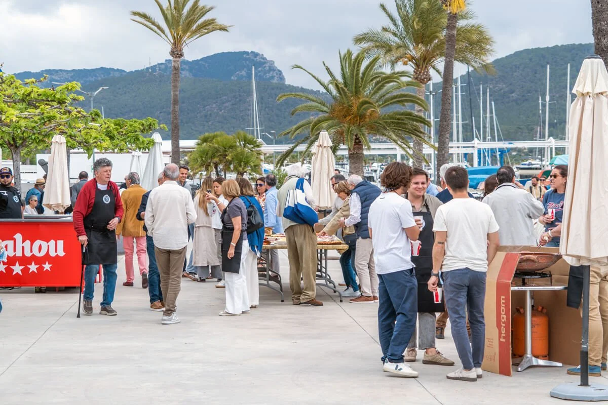 Personas disfrutando de una comida al aire libre junto a un muelle con yates, palmeras y montañas de fondo