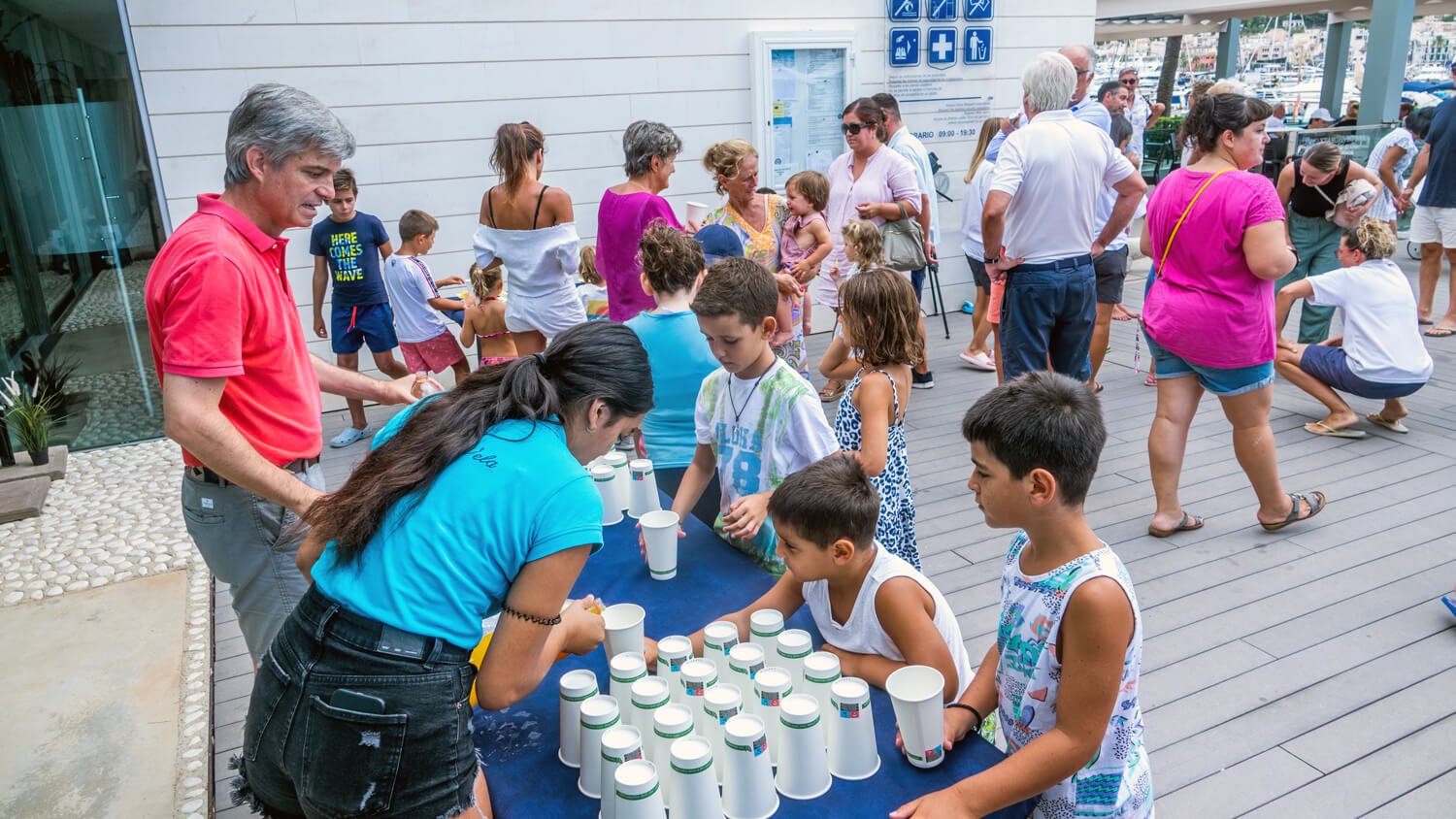 Niños y adultos participando en una actividad al aire libre, con una mesa con vasos desechables, en un evento social o celebración junto a un muelle o zona de embarque.