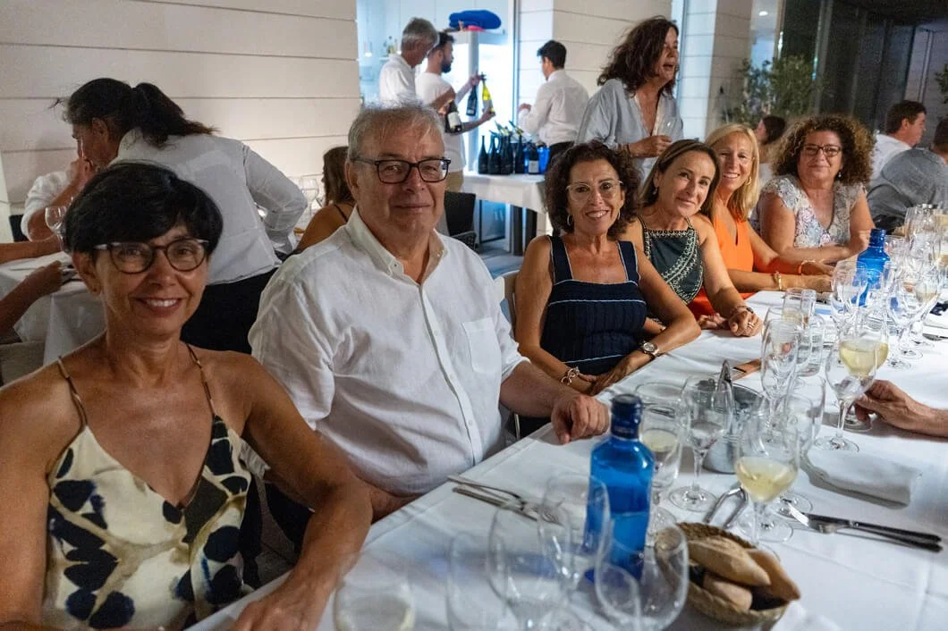 Grupo de personas sentadas en una mesa de comedor en un restaurante, disfrutando de una comida formal, con copas de vino y agua, en un ambiente elegante y luminoso.