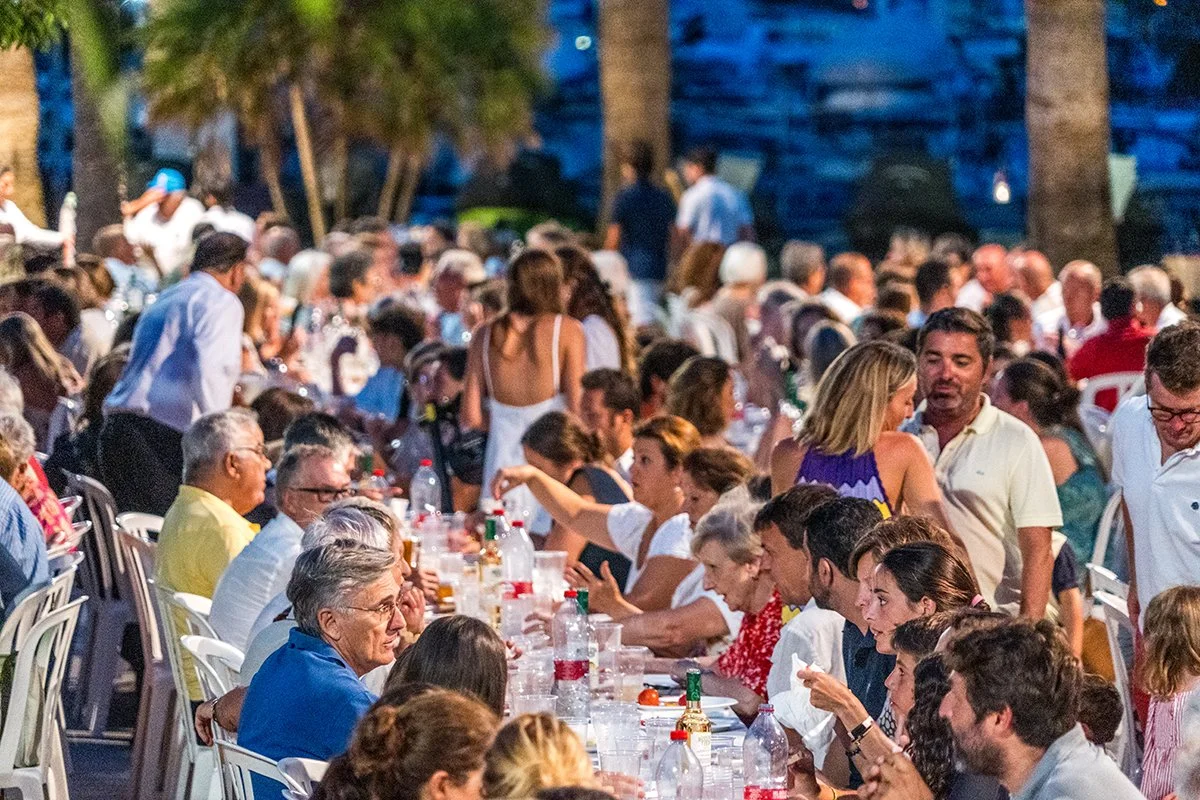 Una gran reunión al aire libre con personas sentadas en una larga mesa, cenando y socializando en un entorno natural con árboles y luz del atardecer.