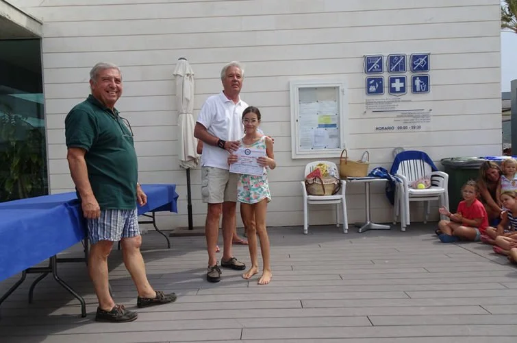Una niña recibe un certificado en un evento al aire libre, con adultos y niños en el fondo, en un área con piso de madera y señalización de emergencias en la pared.